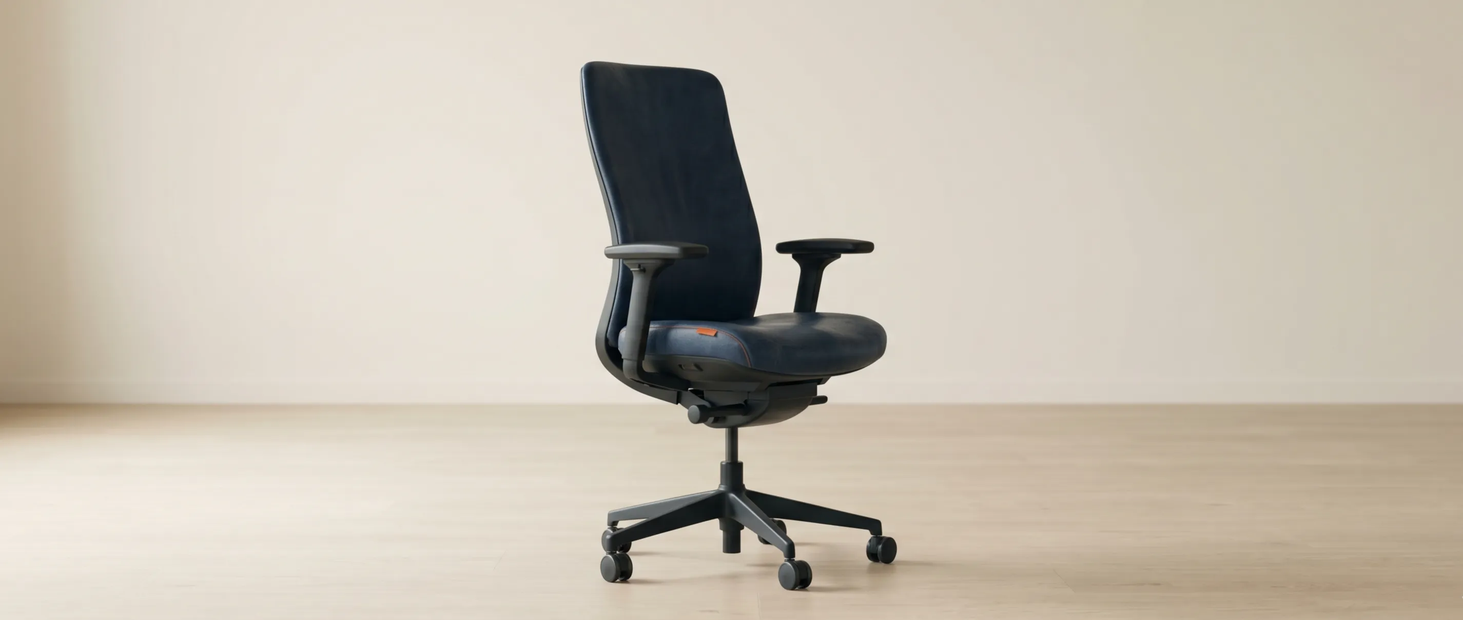 A single well-used office chair standing alone in a quiet, light-filled workspace — the one seat that has been every seat — with a lone burnt-orange detail the only accent.